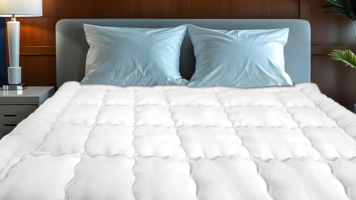 White mattress pad on a bed with light blue pillows and a wooden headboard.